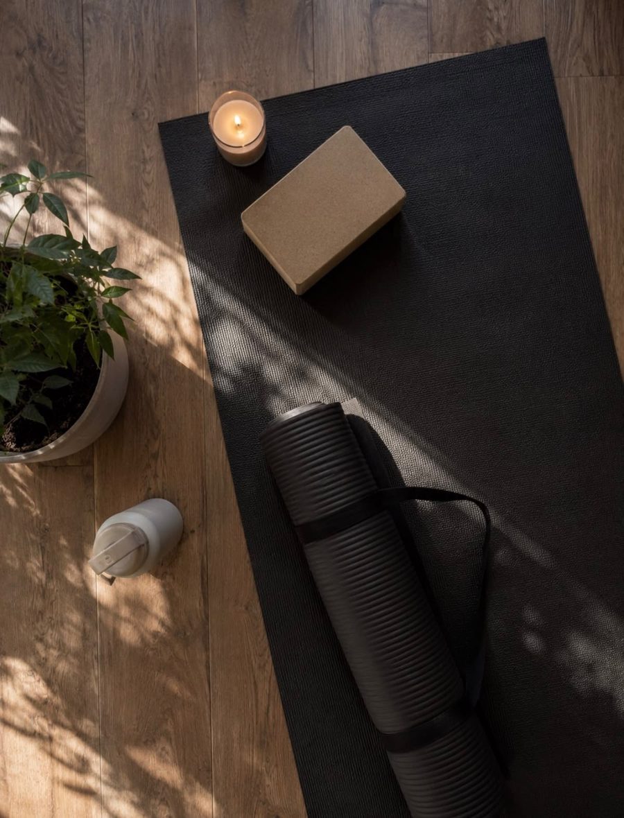 A black yoga mat on warm oak floorboards with a cork block, a lit candle, a water bottle and a plant, in soft dappled sunlight.